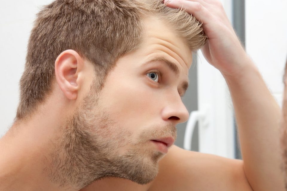 مردی با موهای بلوند مقابل آینه ریزش موی خود را بررسی می کند - درمان ریزش مو در طب سنتی